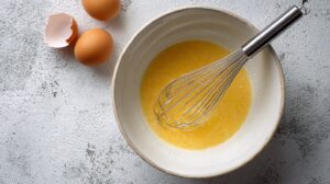 Eggs and melted mixture being whisked in a bowl with cracked eggshells nearby