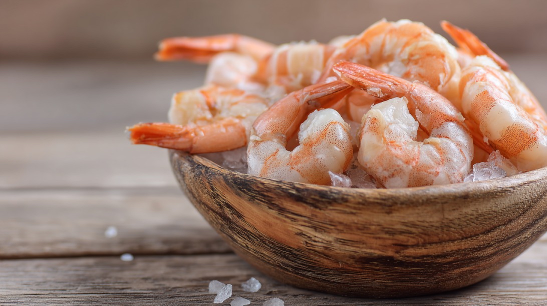 Close up of pink cooked shrimp without shells resting on ice in a wooden bowl on a wooden table