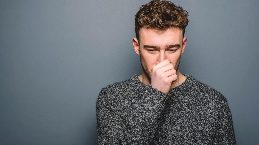 Young man in a gray sweater covering his nose with his hand against a plain background