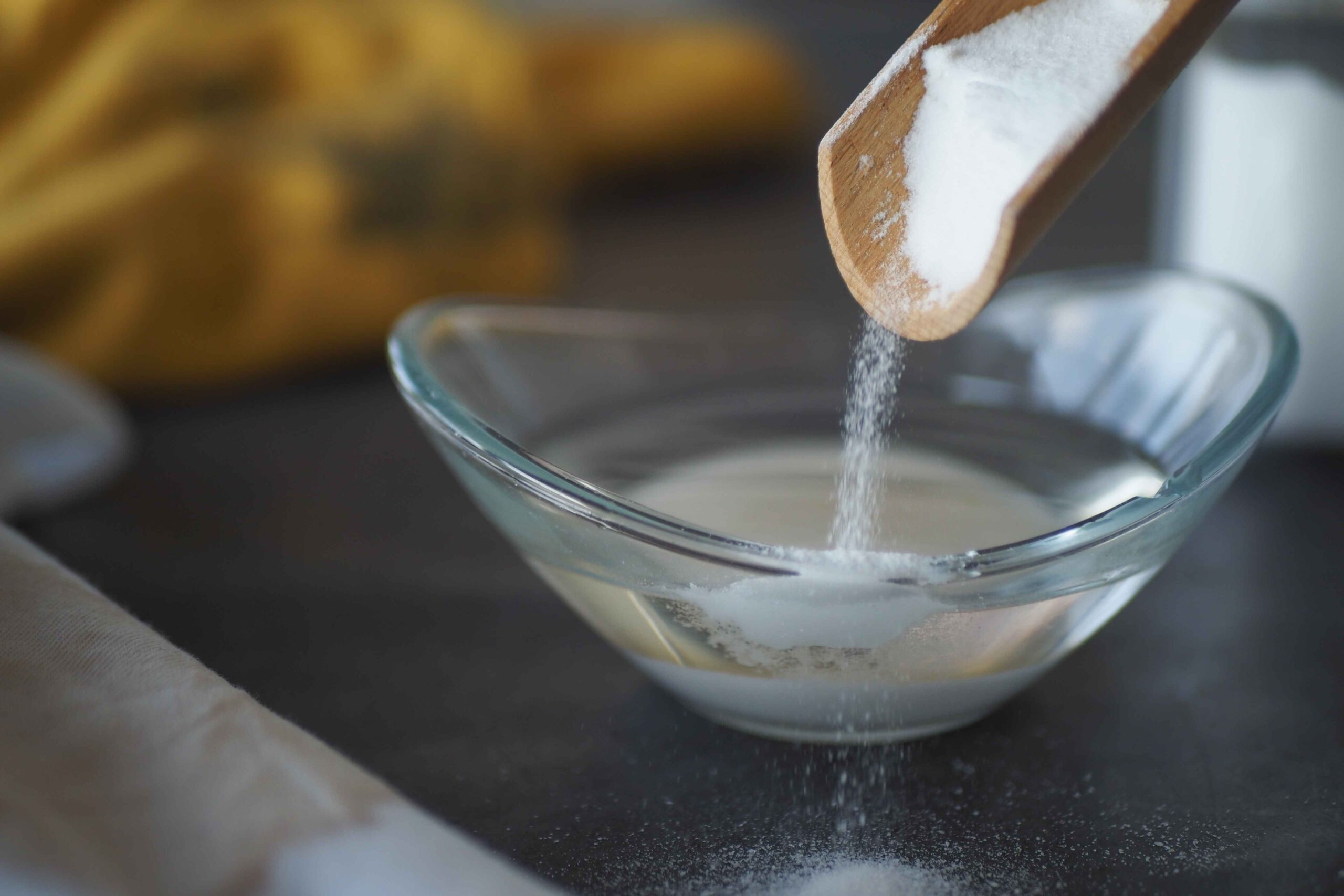 Granulated white baking ingredient being poured from a wooden scoop into a small glass bowl