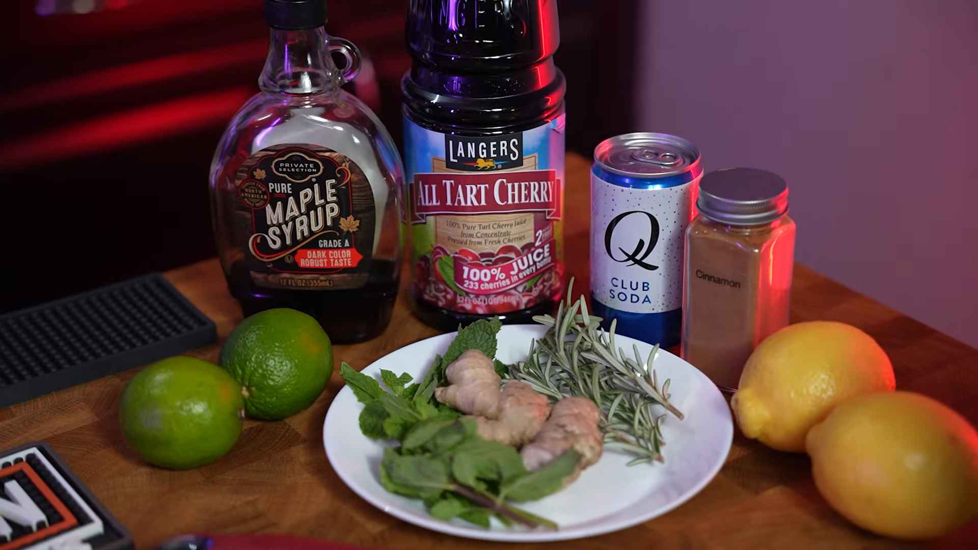 Maple syrup, tart cherry juice, club soda, cinnamon, fresh ginger, mint, rosemary, limes, and lemons arranged on a countertop