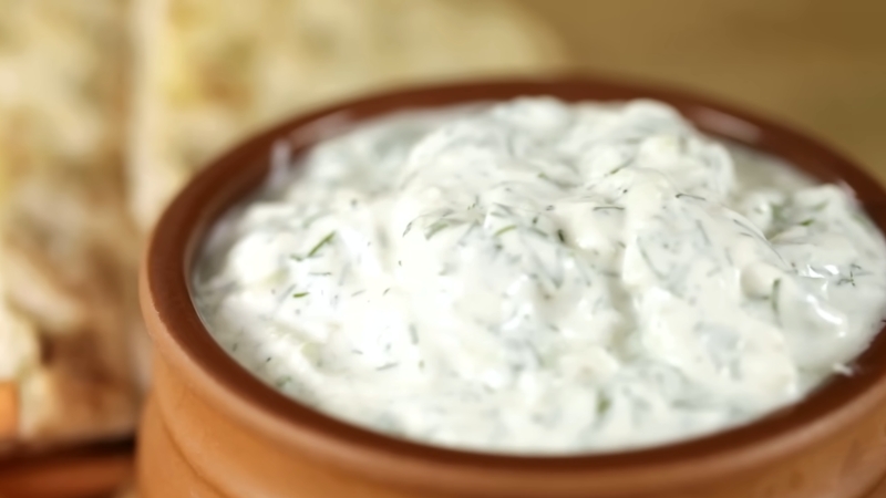 A bowl of creamy tzatziki sauce with visible cucumber and dill, served with flatbread in the background