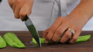 A person slicing a peeled cucumber on a cutting board with a knife