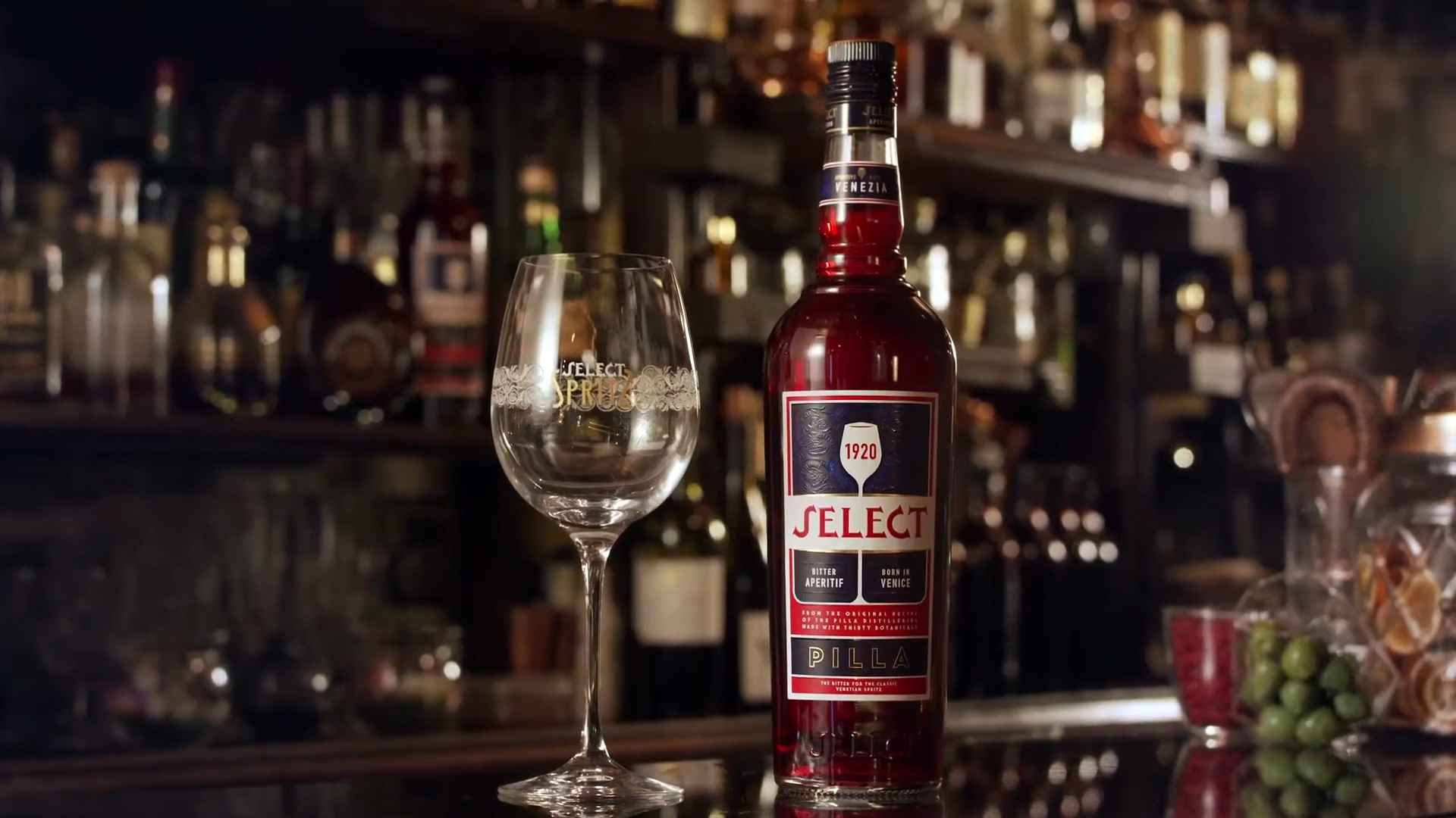 Bottle of Select Aperitivo on a bar counter beside a branded wine glass, with liquor bottles blurred in the background
