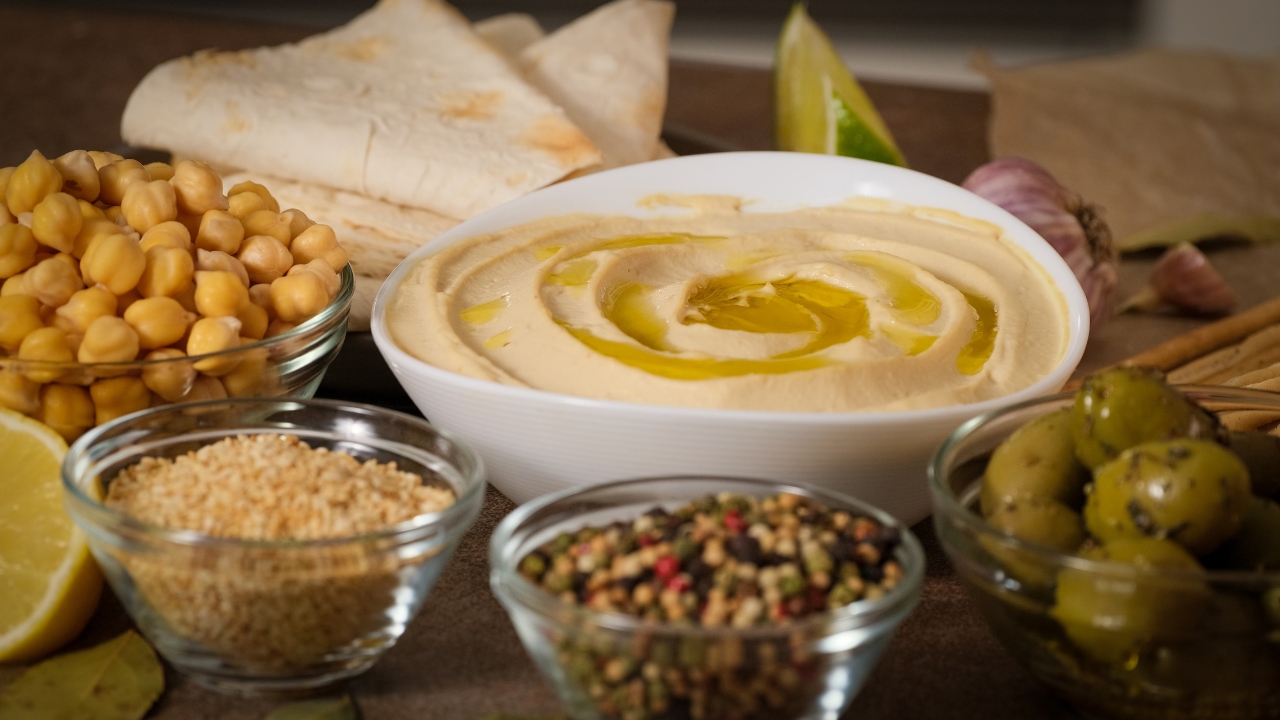 Bowl of hummus with olive oil surrounded by chickpeas, sesame seeds, olives, and pita bread