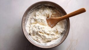 Bowl of cottage cheese dough being mixed with flour using a wooden spoon