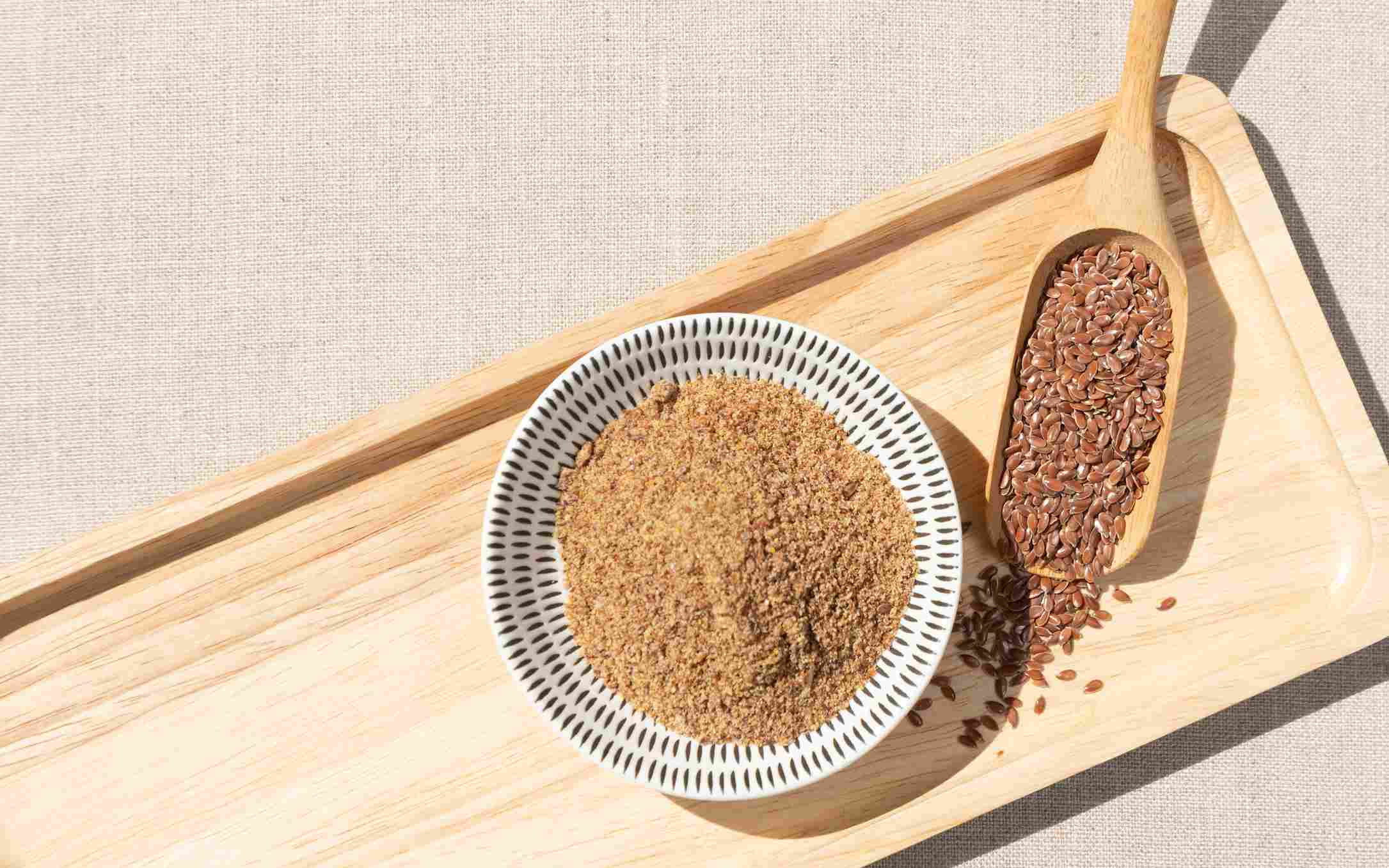 Ground flaxseed in a bowl with whole flaxseeds in a wooden scoop on a tray
