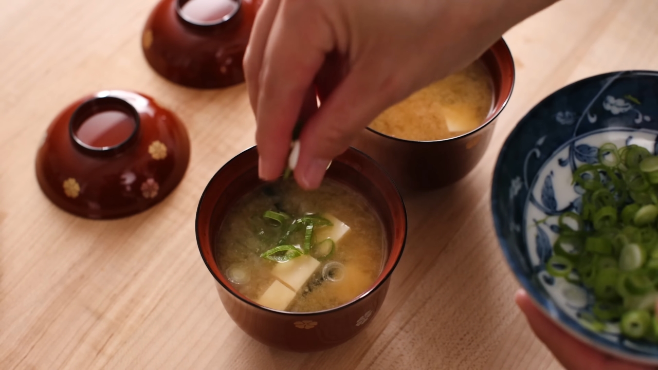 Hand adding green onions to a bowl of miso soup with tofu and seaweed