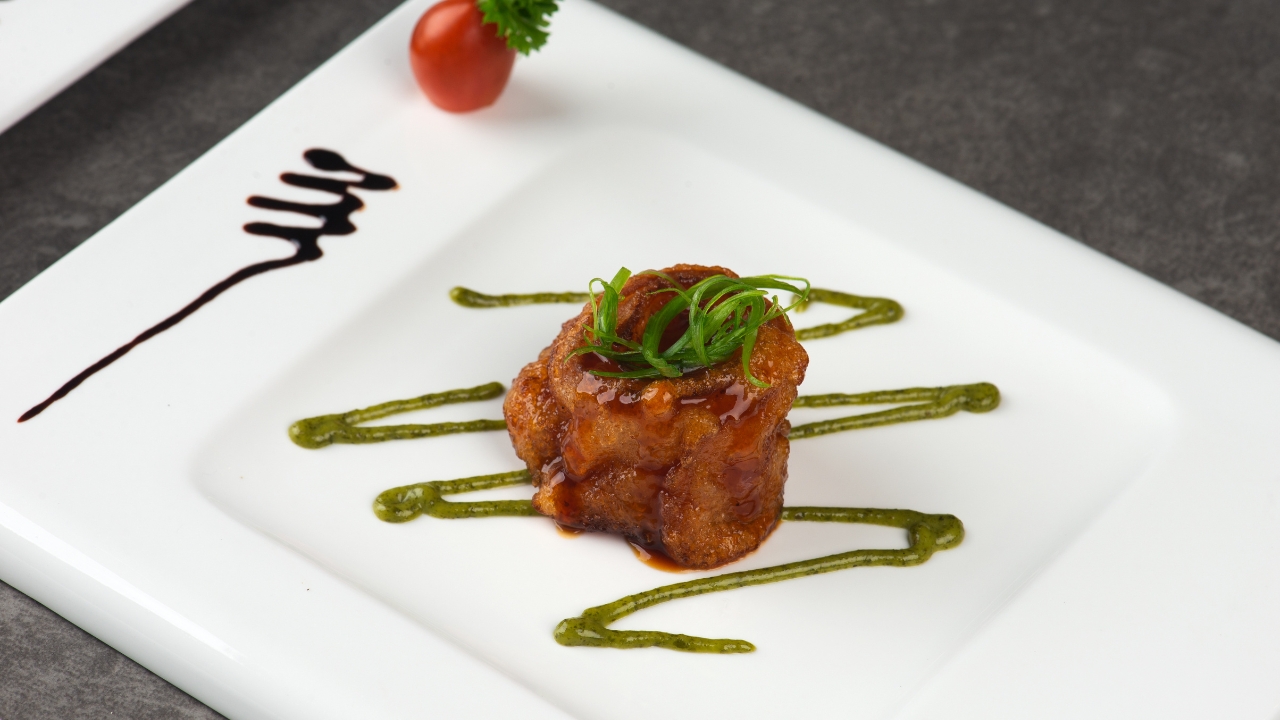 A small glazed bite sits on a white plate with green sauce lines and a dark sauce mark
