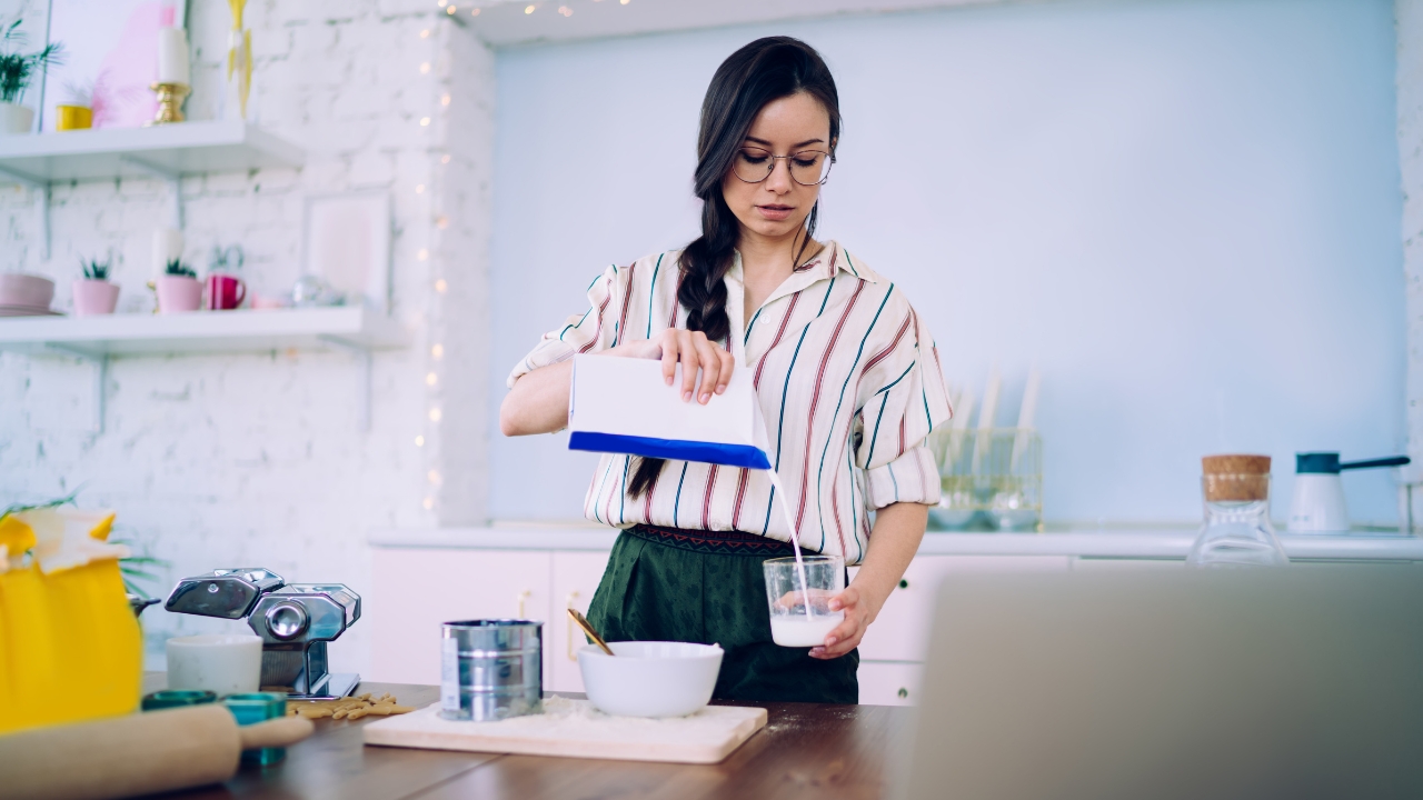 Woman pours milk into a glass cup while preparing ingredients in a kitchen