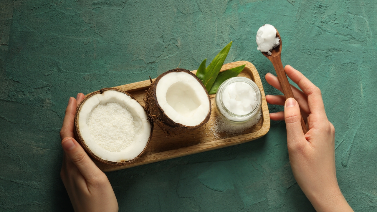 Hands hold coconut pieces, coconut flakes, and a jar of coconut oil on a wooden tray