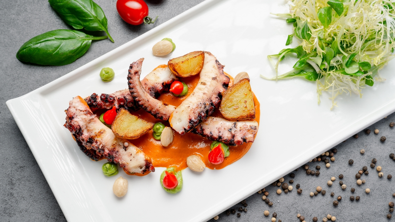 Grilled octopus plated with sauce, potatoes, greens, and herbs on a white rectangular plate