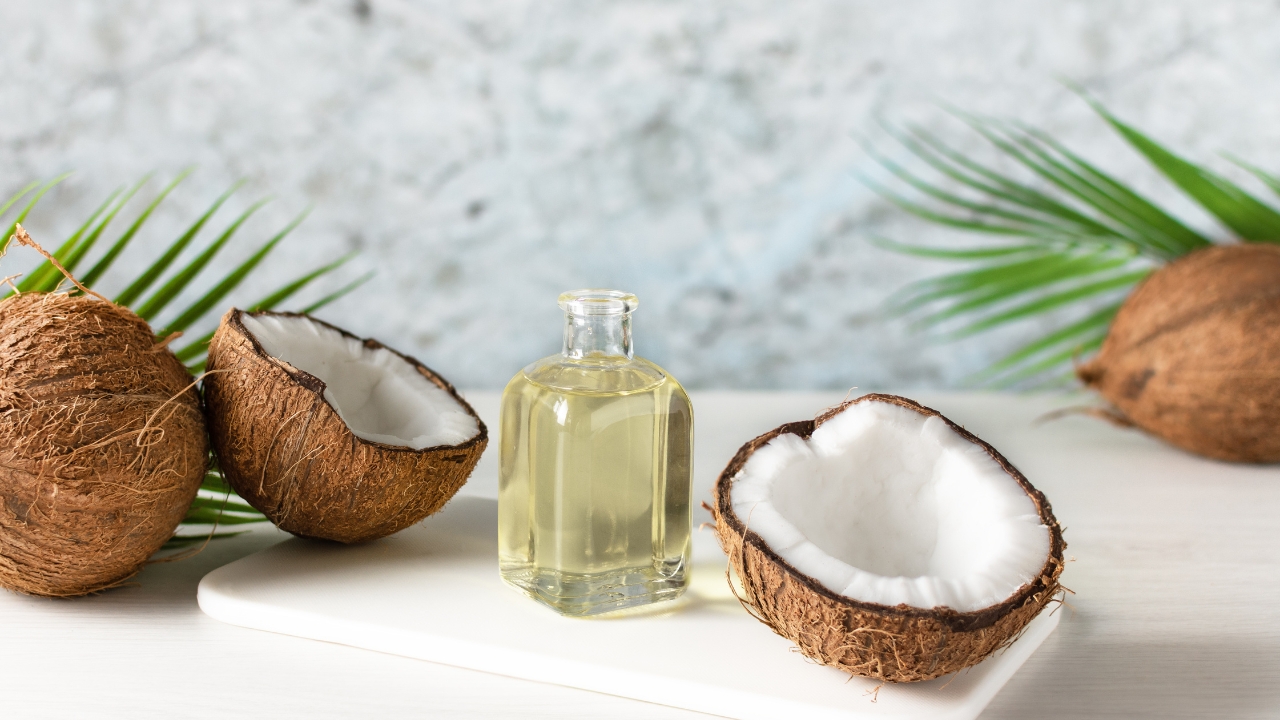 A glass bottle of coconut oil sits between fresh coconut halves on a light table