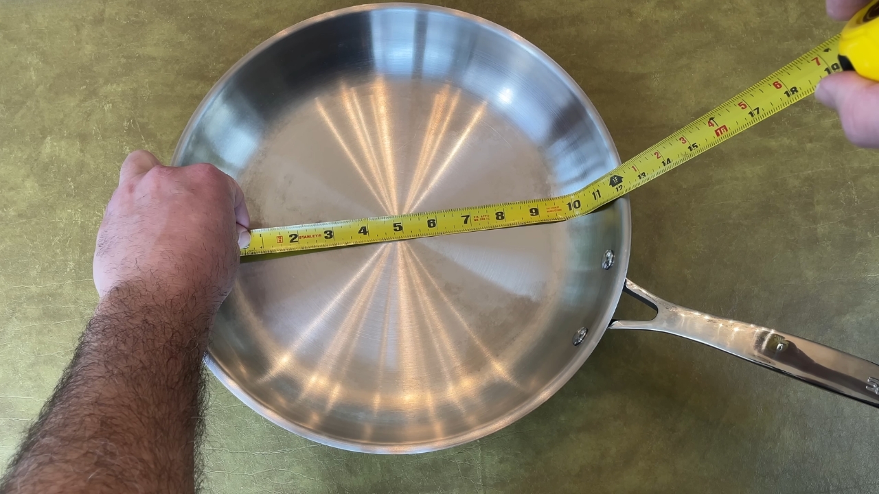 A stainless steel skillet is measured across the top with a yellow tape measure