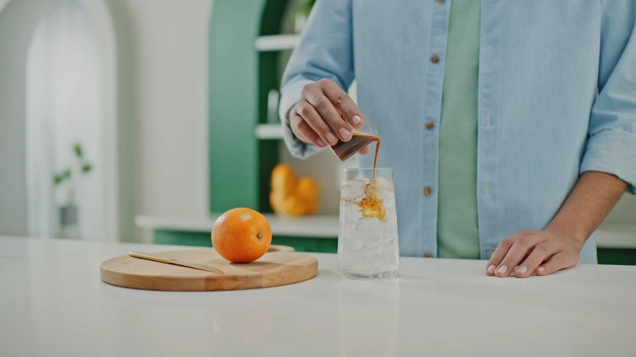 A person adds espresso to a glass of tonic water with ice beside an orange