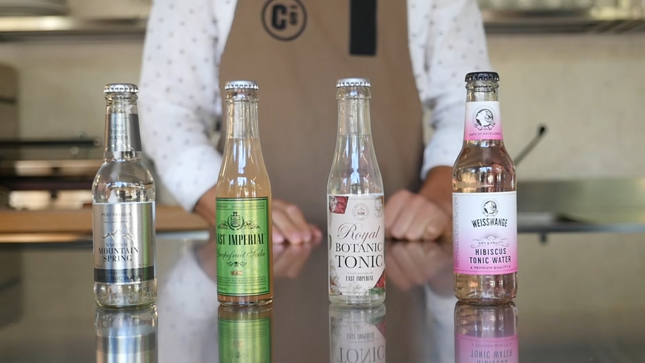 Four bottles of tonic water sit on a counter beside a barista