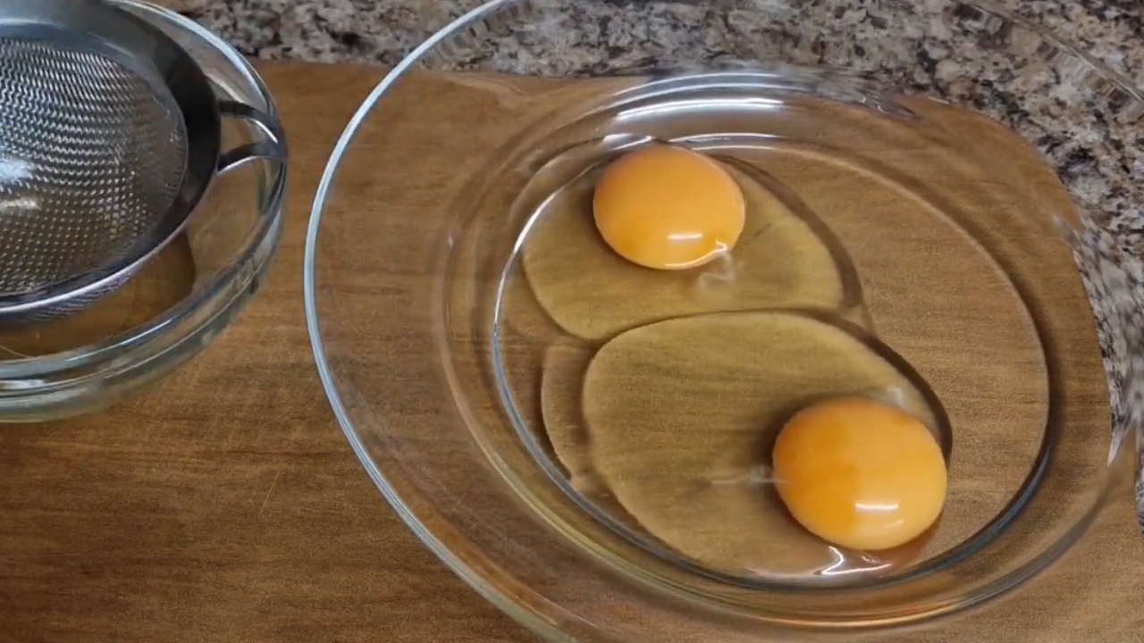 Two raw eggs cracked into a glass bowl beside a fine-mesh strainer.